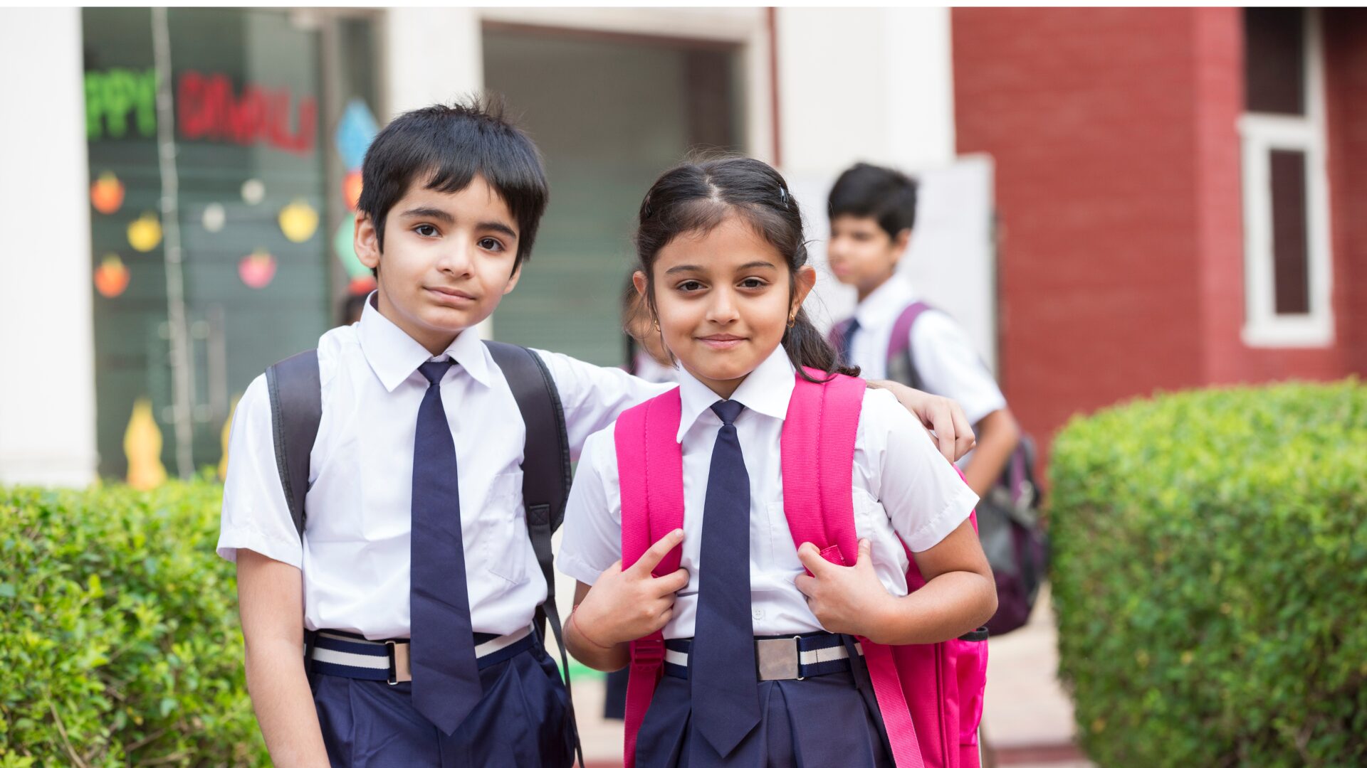 Two South Asian children in school uniforms standing in front of a school building, smiling at the camera — representing Telugu children in diaspora learning environments